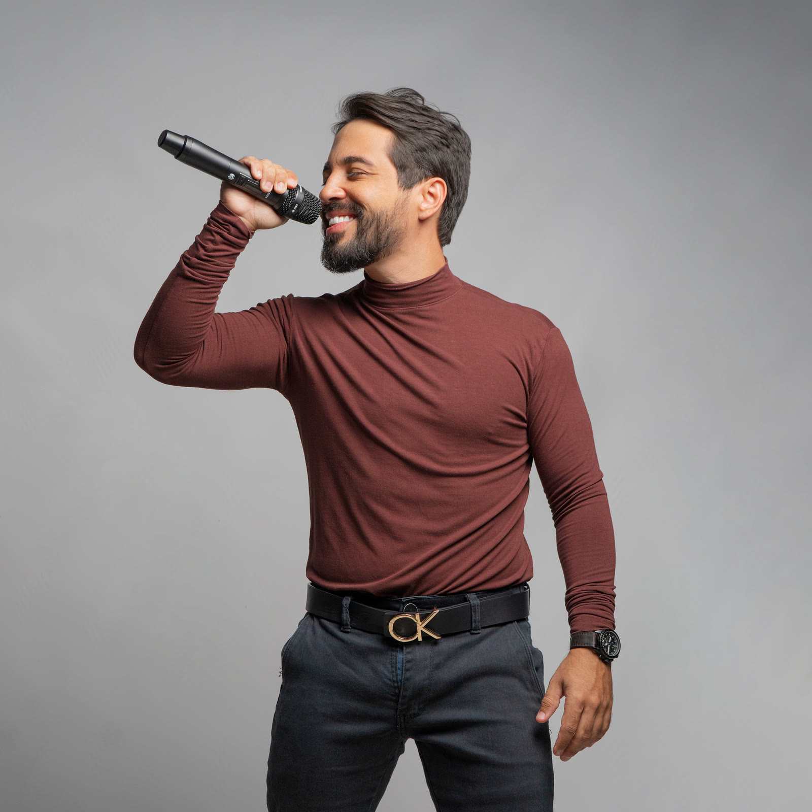 Brener Coelho sorrindo enquanto canta ao microfone, de blusa bordô